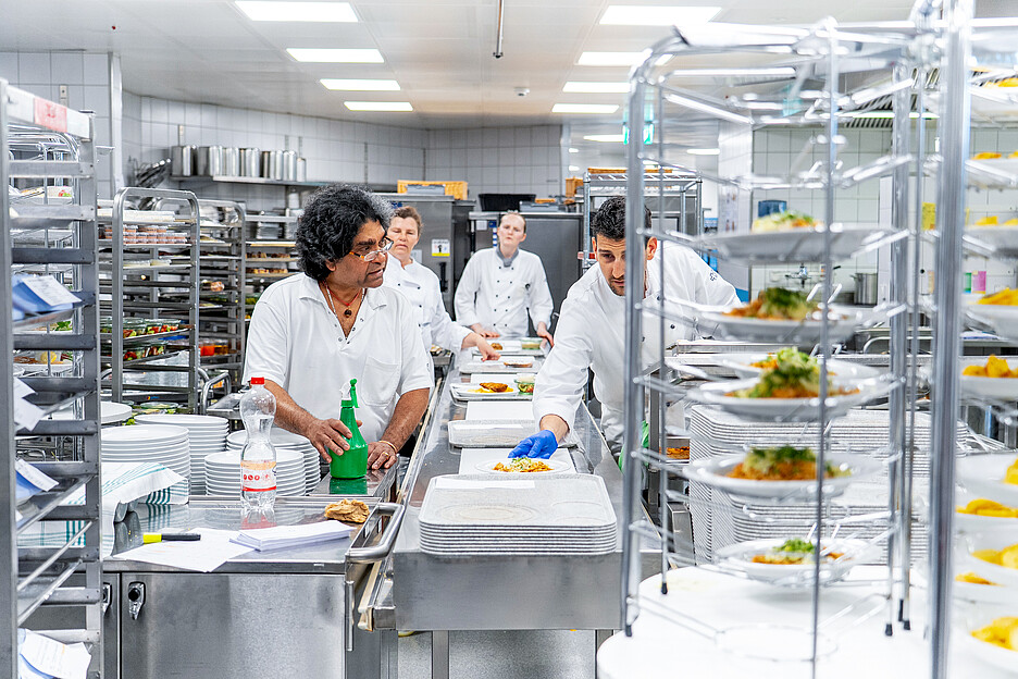 Les plats froids et chauds dressés sont positionnés dans de la vaisselle prévue à cet effet sur la bande centrale de distribution de repas de Schmalz Distributions-Systeme AG sur le plateau E-Norm.